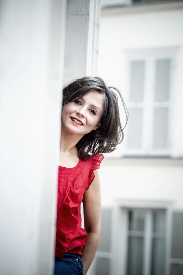Nadia Battiston en haut rouge, souriante, devant une façade aux volets blancs
